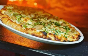 Freshly baked wood-fired pizza with bell peppers, olives, and melting cheese placed on a wooden tray under the open sky.
