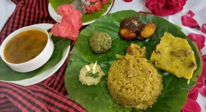 A colorful tribal thali with rice, dal, bamboo shoot curry, and local pickles served on a wooden table with a green forest view.